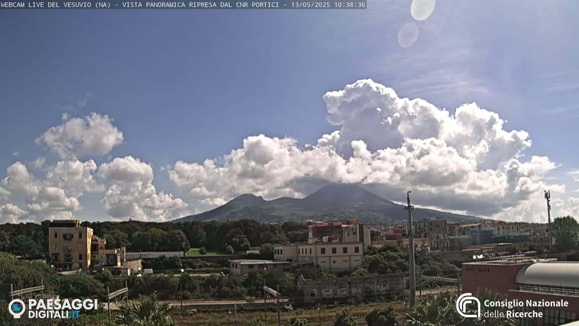 A partly cloudy sky reveals Mount Vesuvius in the distance, overlooking a town with buildings and green spaces.