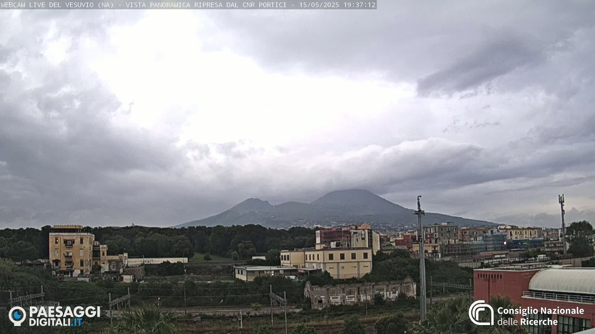 A view of Mount Vesuvius in the distance, under a cloudy sky, with buildings and greenery in the foreground.
