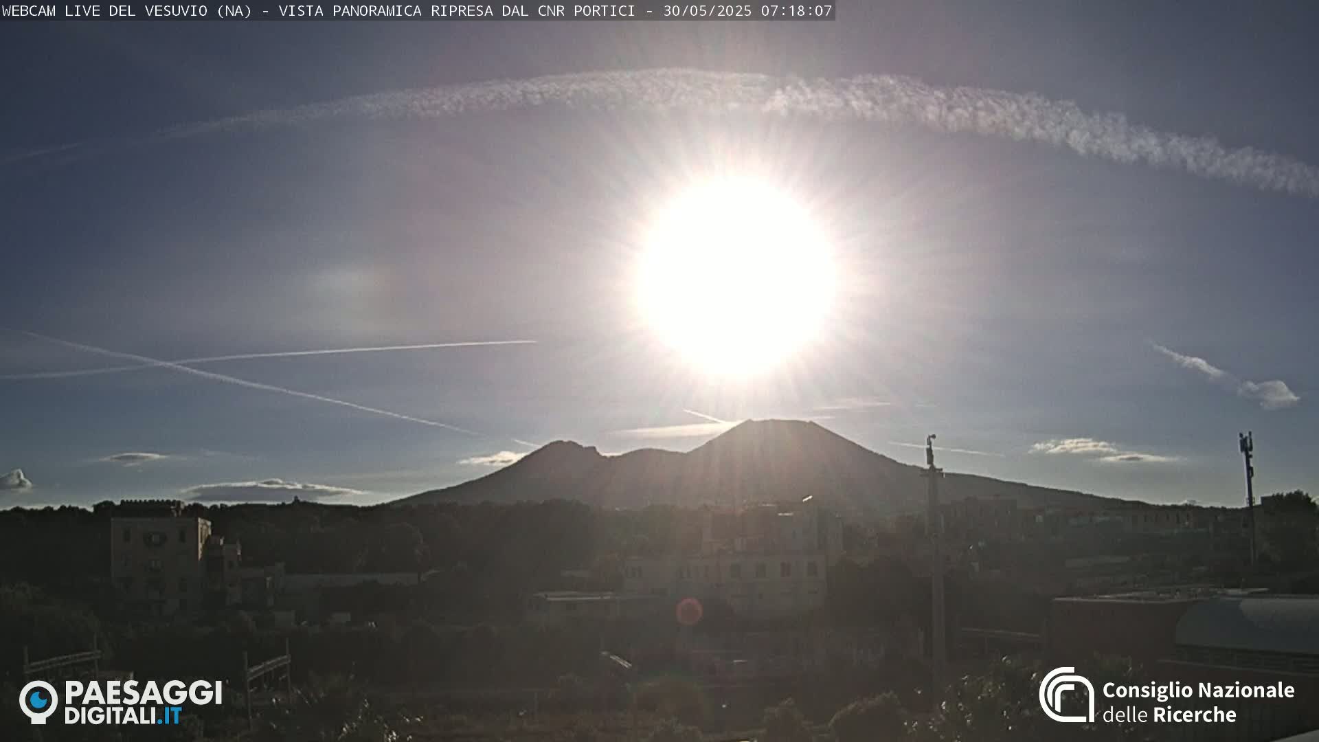 A bright sun shines over a distant, dark mountain range and a town, with a few contrails visible in the mostly clear sky.