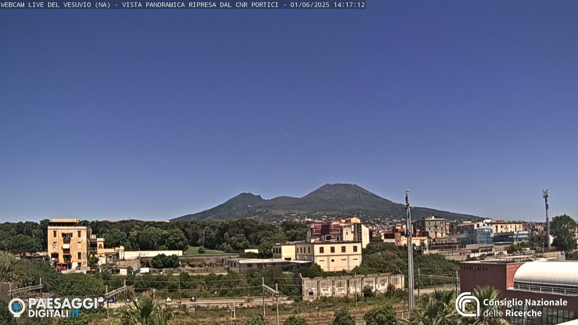 A view of Mount Vesuvius in the distance, under a clear blue sky, with buildings and greenery in the foreground.