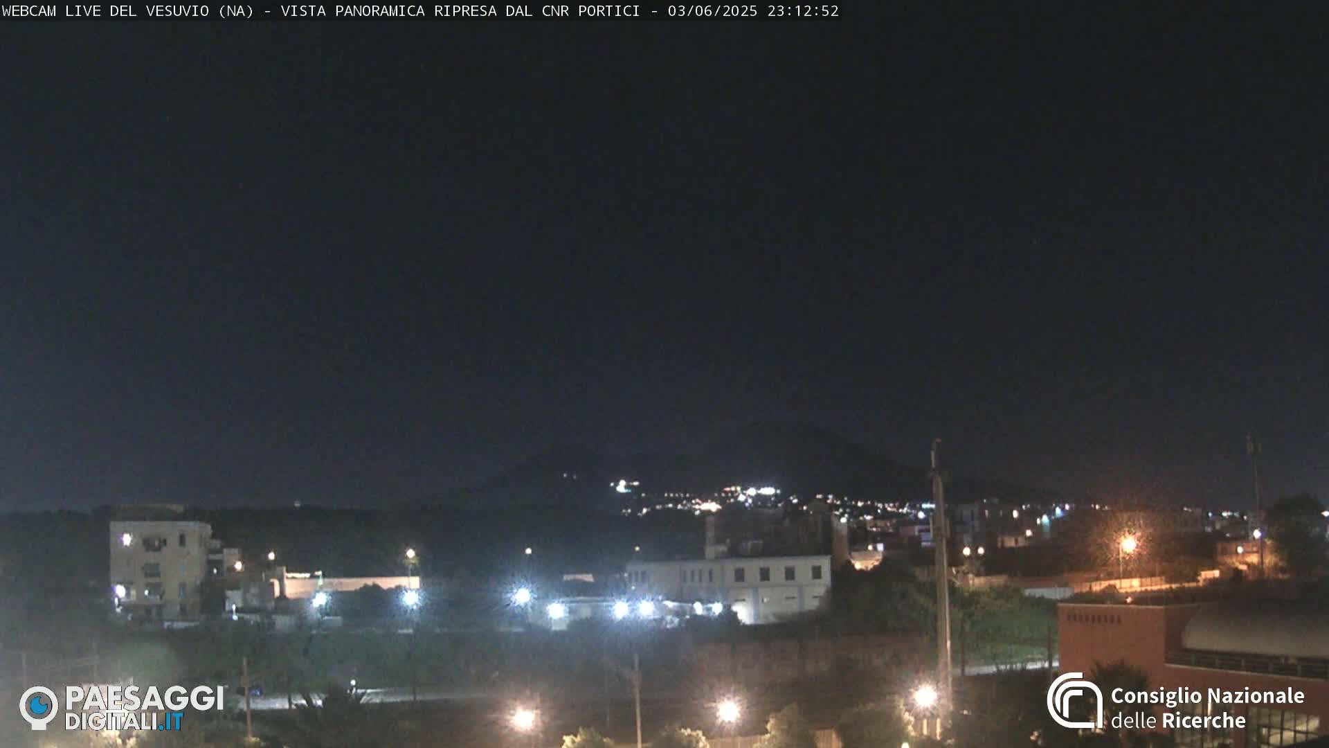 A nighttime long shot of a town with scattered lights, a hill in the background, and a dark, clear sky.