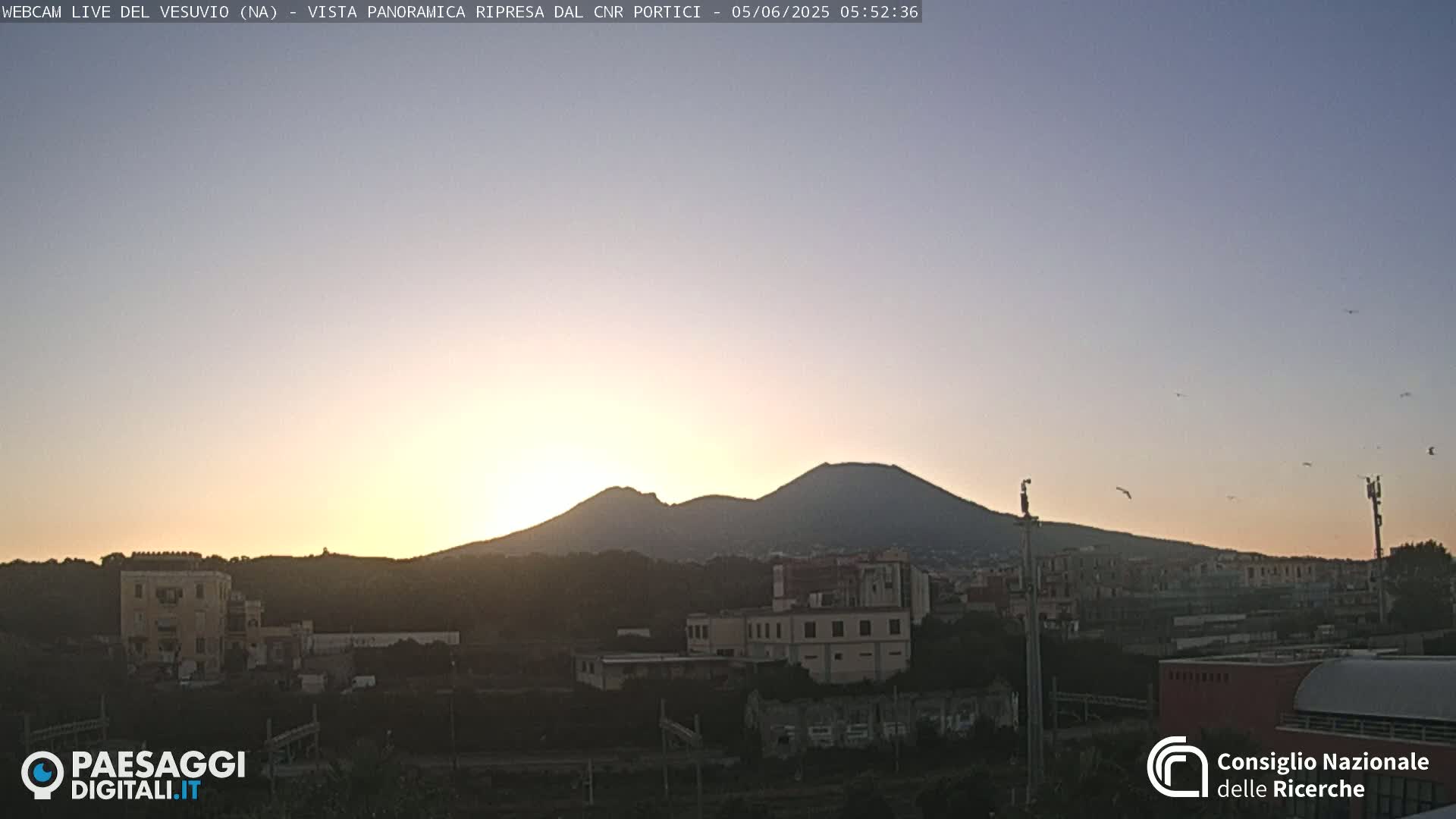 A sunrise casts a warm glow over a cityscape with Mount Vesuvius in the background under a clear sky.