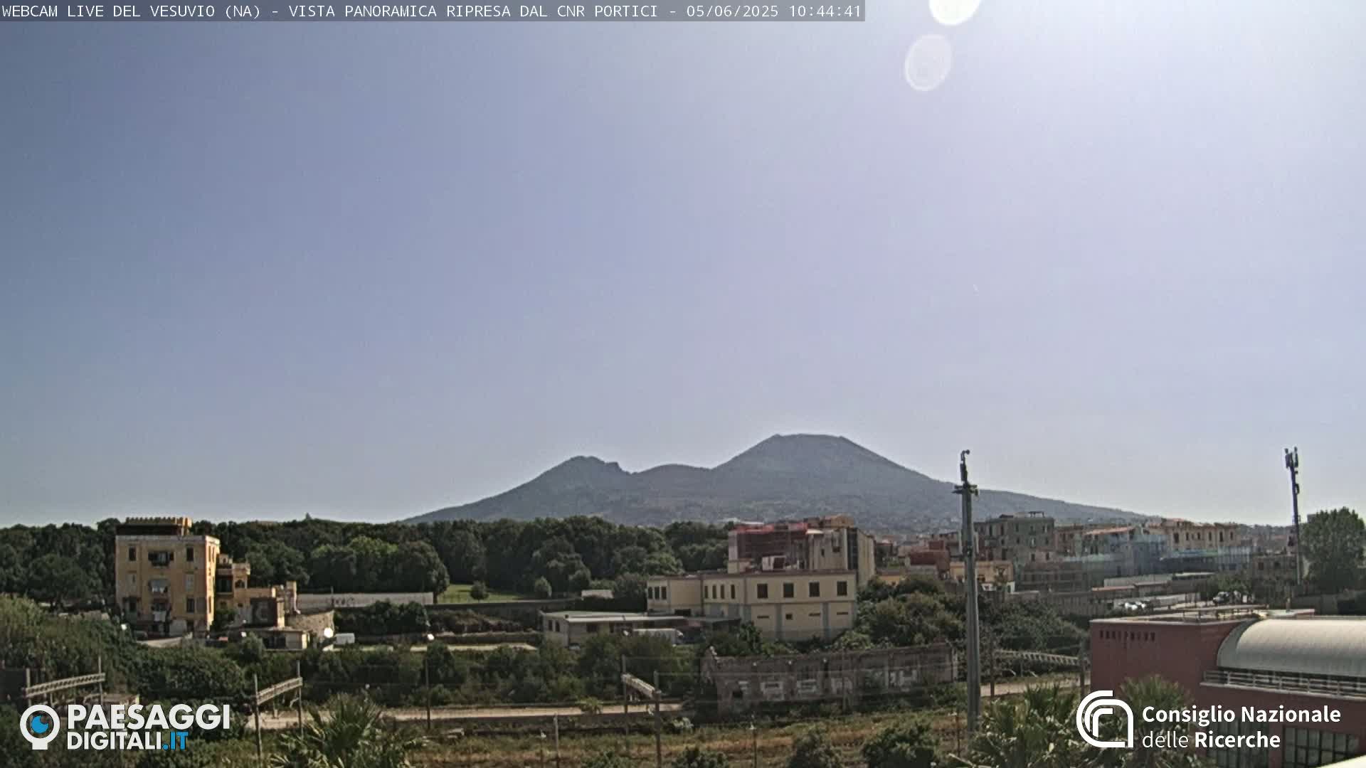 A distant, hazy view of Mount Vesuvius under a clear, light-blue sky, with buildings and trees in the foreground.