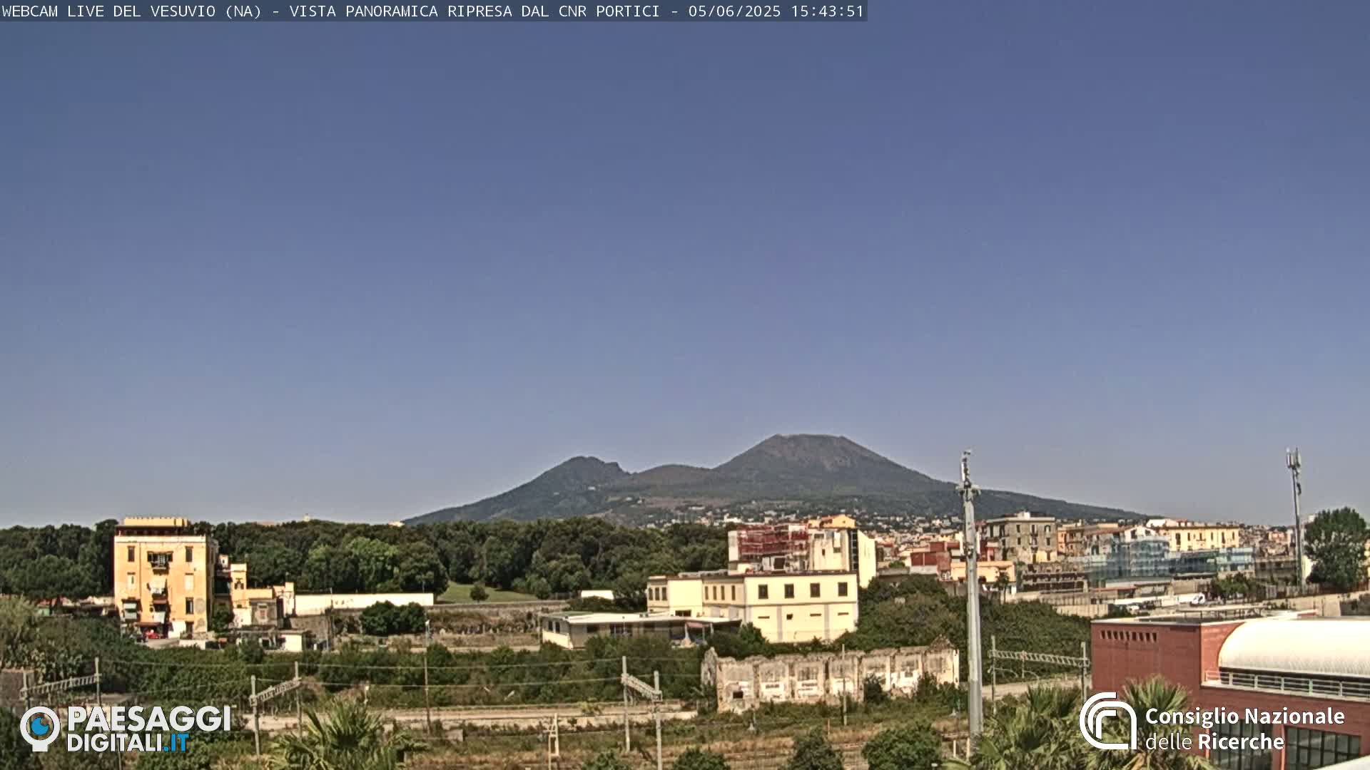 Mount Vesuvius is visible in the distance under a clear blue sky, behind a cityscape featuring low-rise buildings and green vegetation.