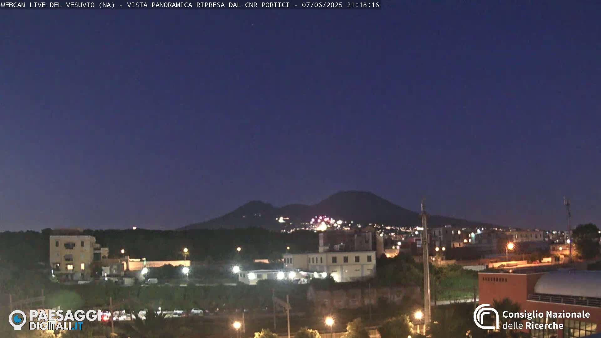 A nighttime view of a city illuminated by streetlights, with a dark, mountainous silhouette in the background under a twilight sky.