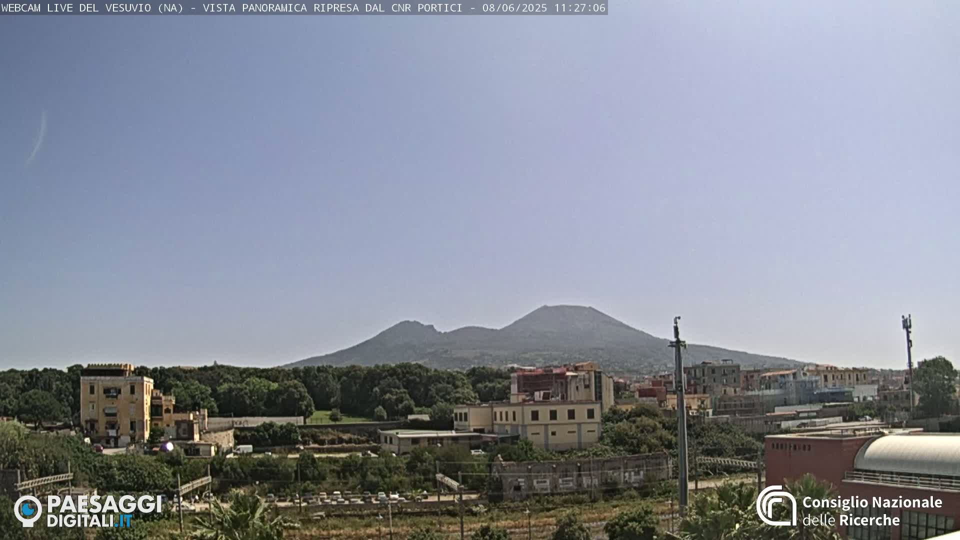 A distant view of Mount Vesuvius on a clear, sunny day, with buildings and trees in the foreground.
