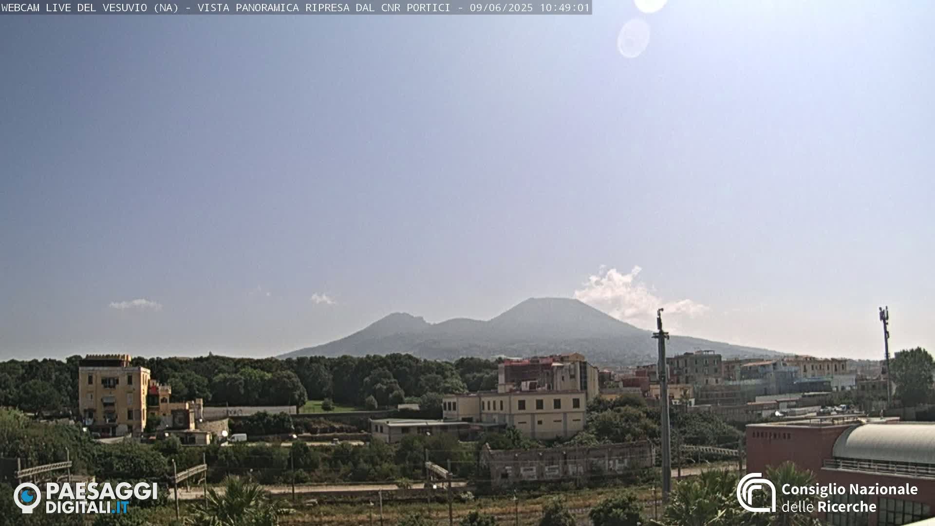 A hazy, sunny day reveals Mount Vesuvius in the distance, overlooking a town with buildings and greenery in the foreground.