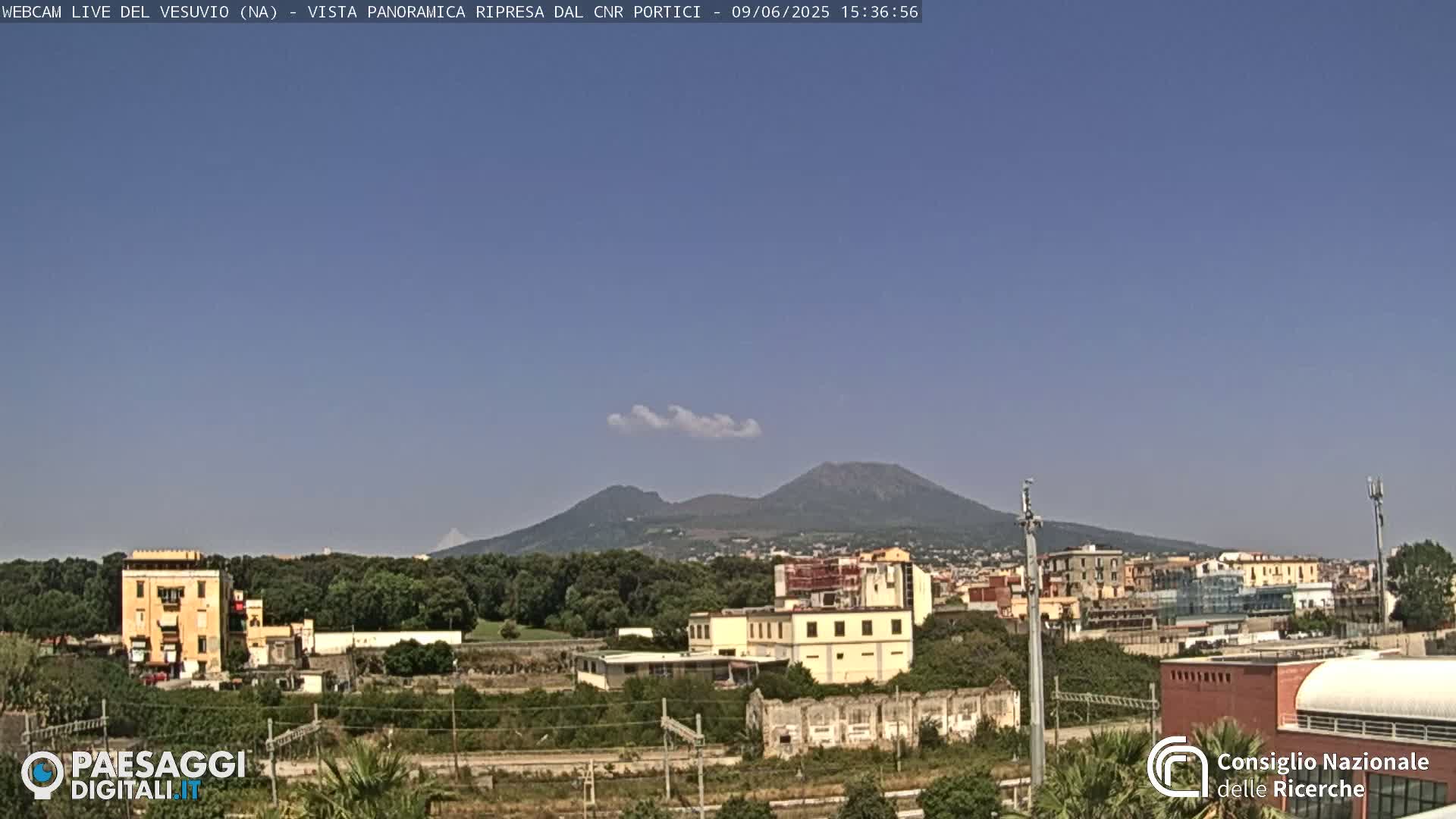 A distant view of Mount Vesuvius under a clear blue sky, with a small, thin cloud above it and a town in the foreground.