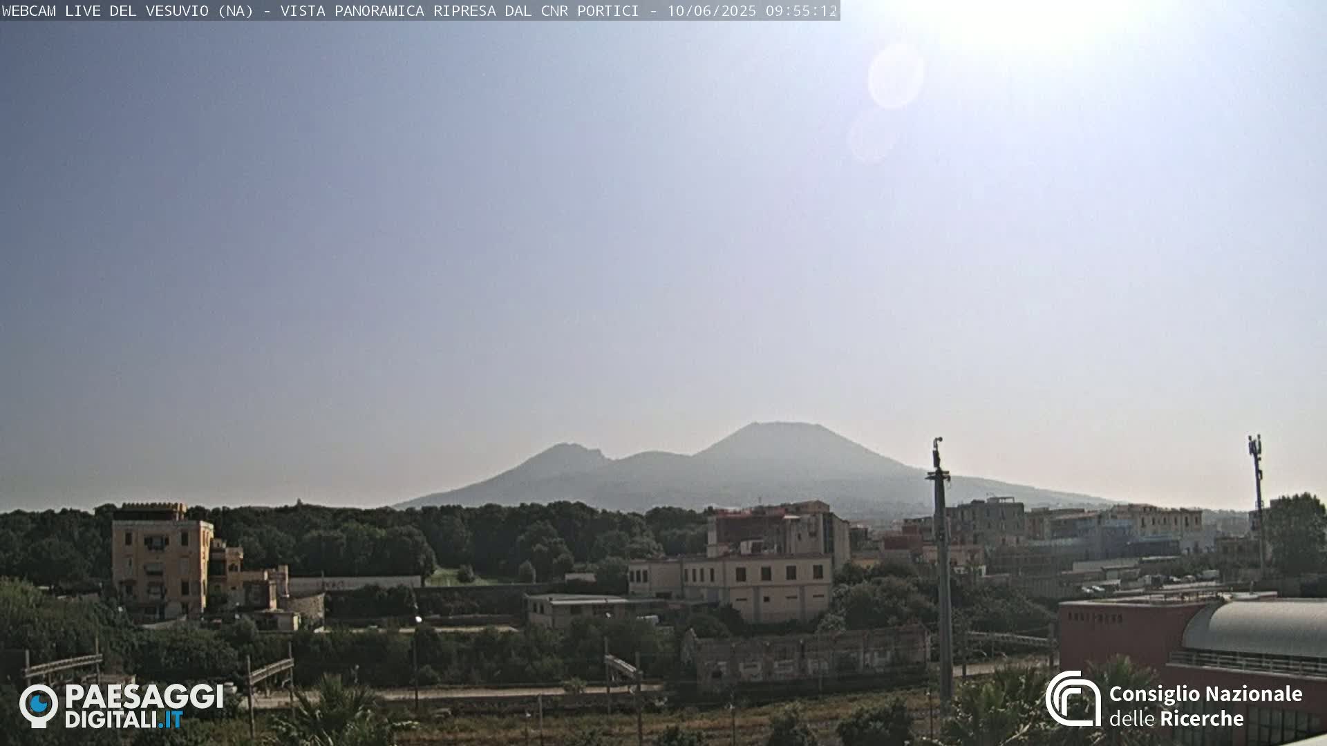 A hazy mountain range is visible in the distance, beyond a town of low-rise buildings and lush green vegetation under a bright, sunny sky.