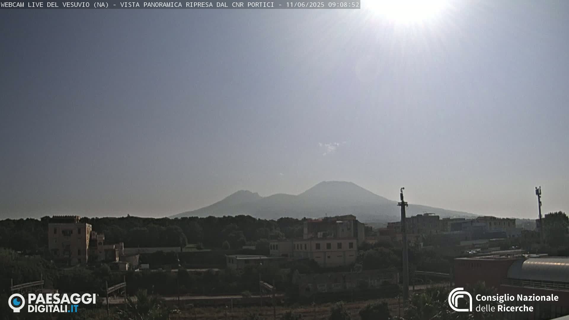A hazy, sunny day reveals Mount Vesuvius in the distance, overlooking a town with low-rise buildings and lush green vegetation.
