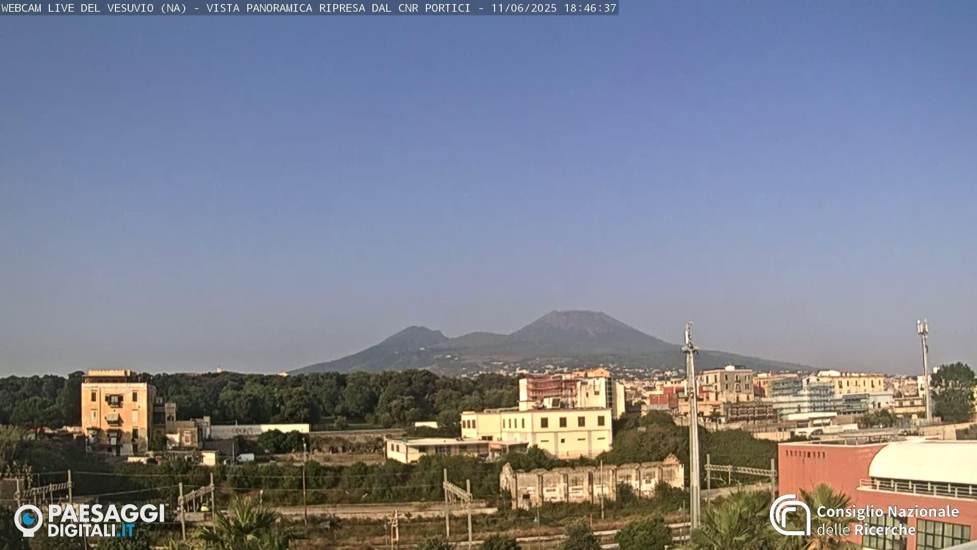 A distant view of Mount Vesuvius under a clear blue sky, overlooking a cityscape with low-rise buildings and some greenery.