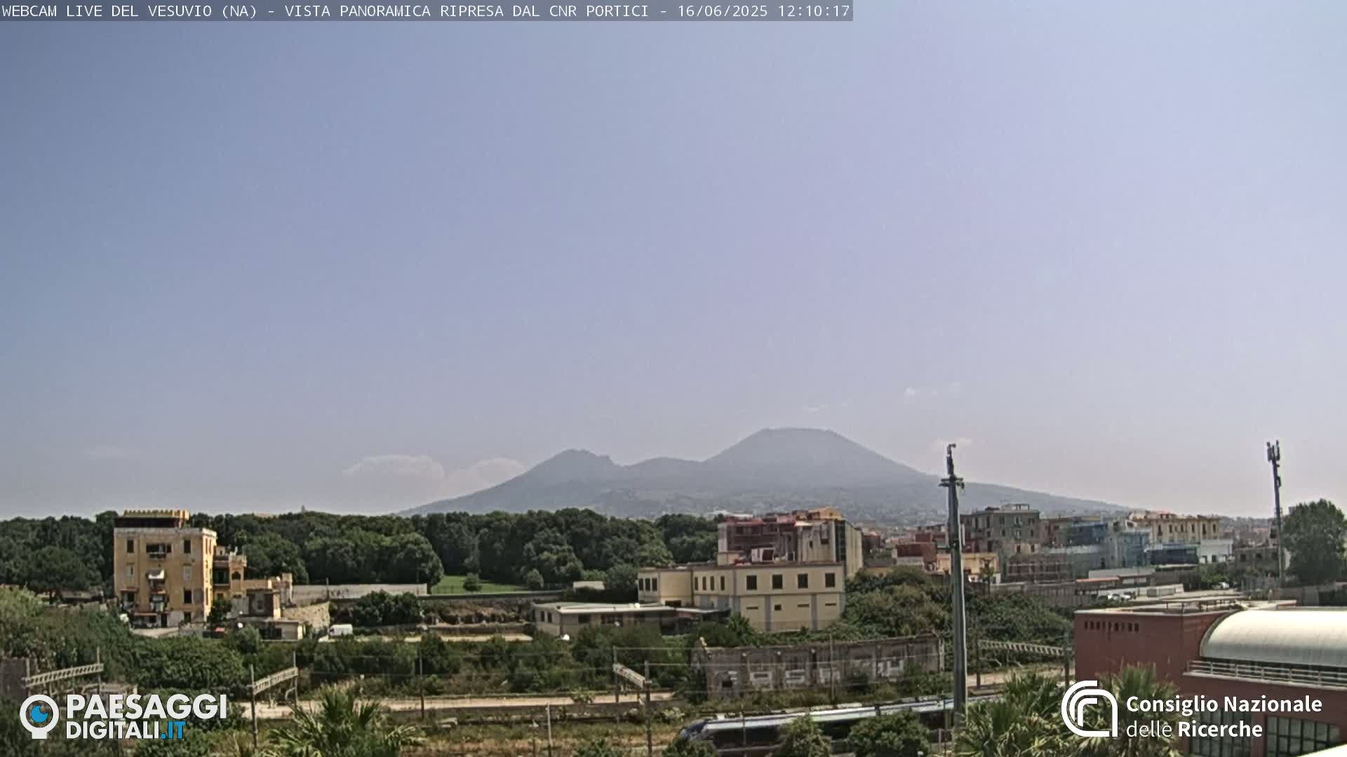 A distant, hazy view of Mount Vesuvius on a clear, sunny day, overlooking a suburban landscape with buildings and trees in the foreground.