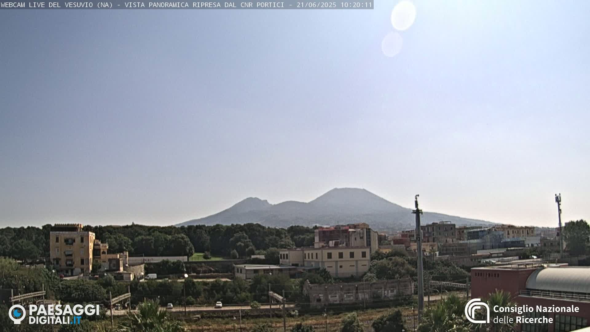 A distant, hazy view of Mount Vesuvius on a clear, sunny day, with buildings and trees in the foreground.