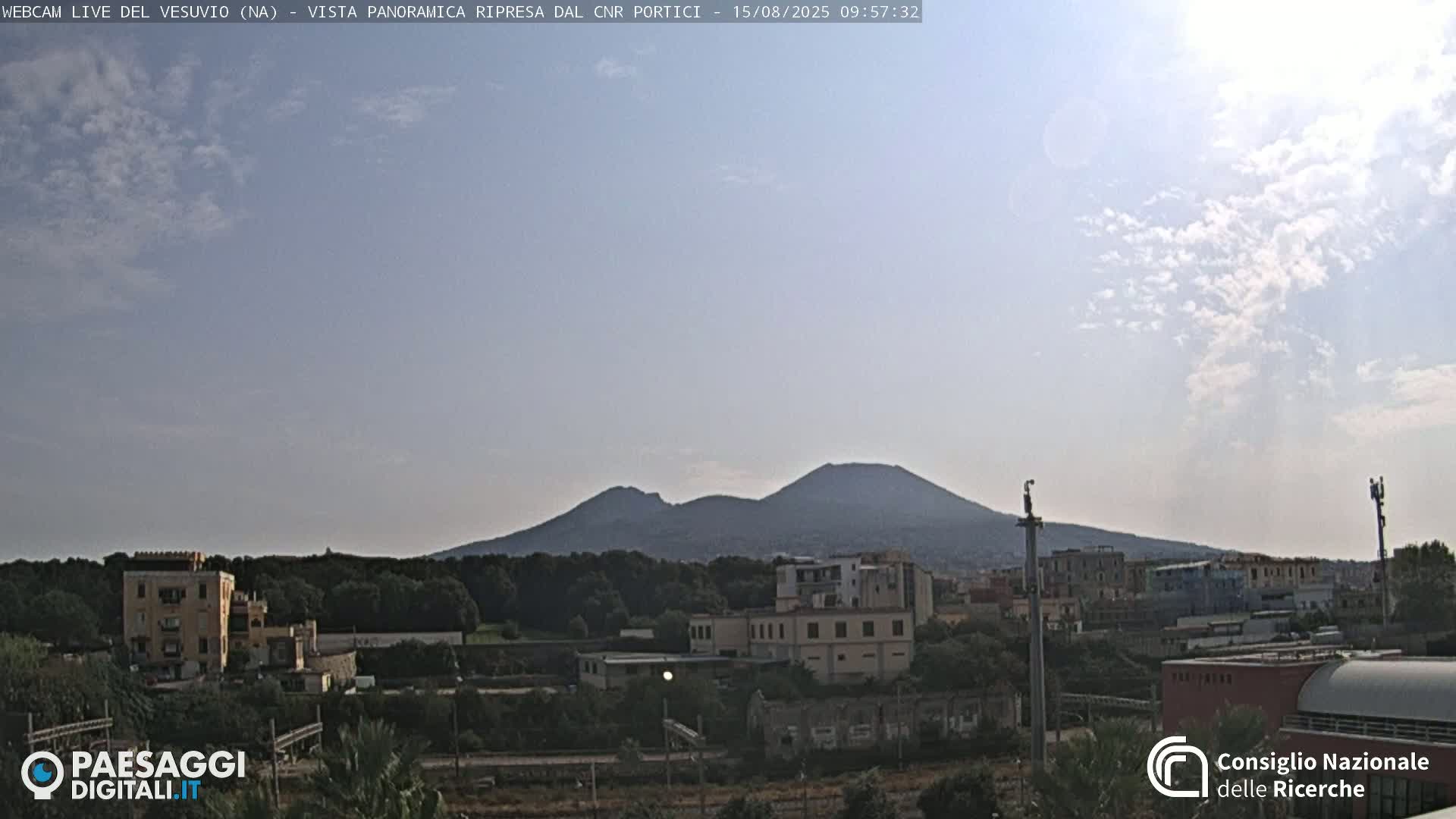 A partly cloudy sky overlooks a cityscape with Mount Vesuvius in the background.