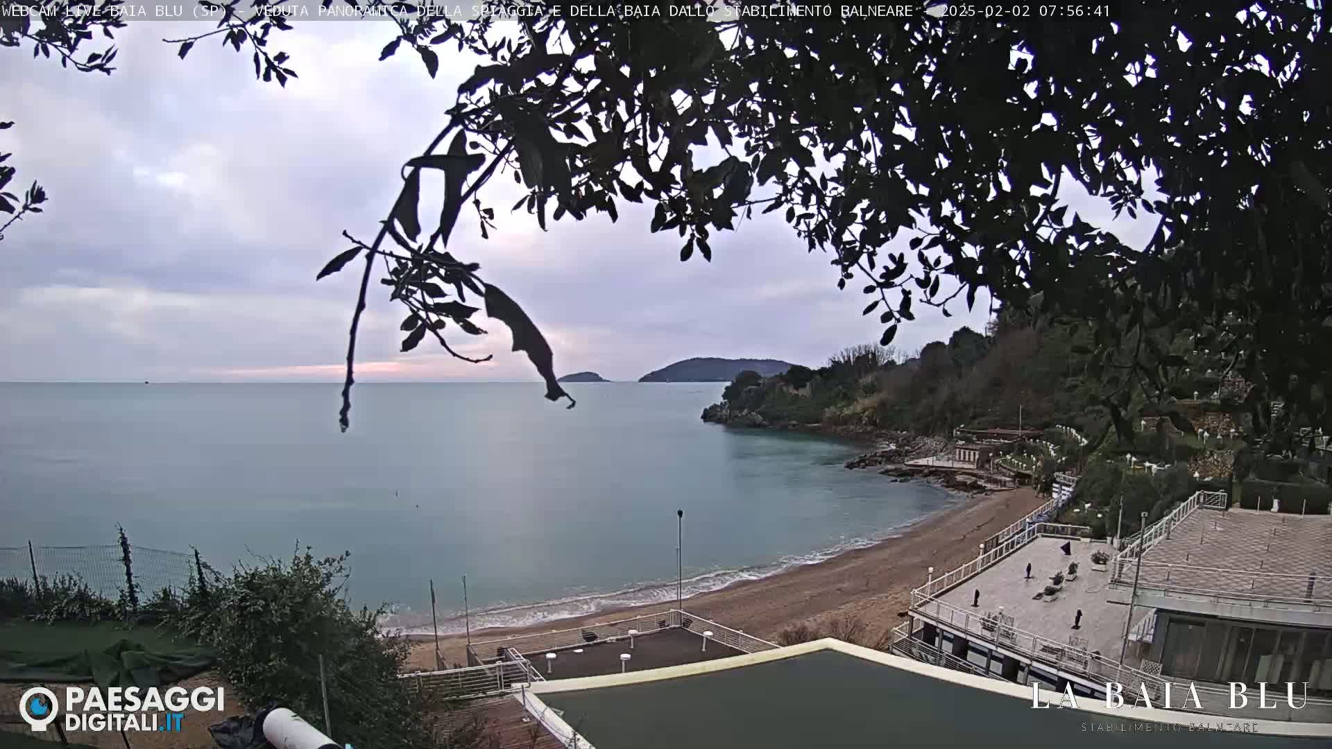 A mostly cloudy day overlooks a calm bay with a sandy beach and buildings; some bare trees partially obscure the view.