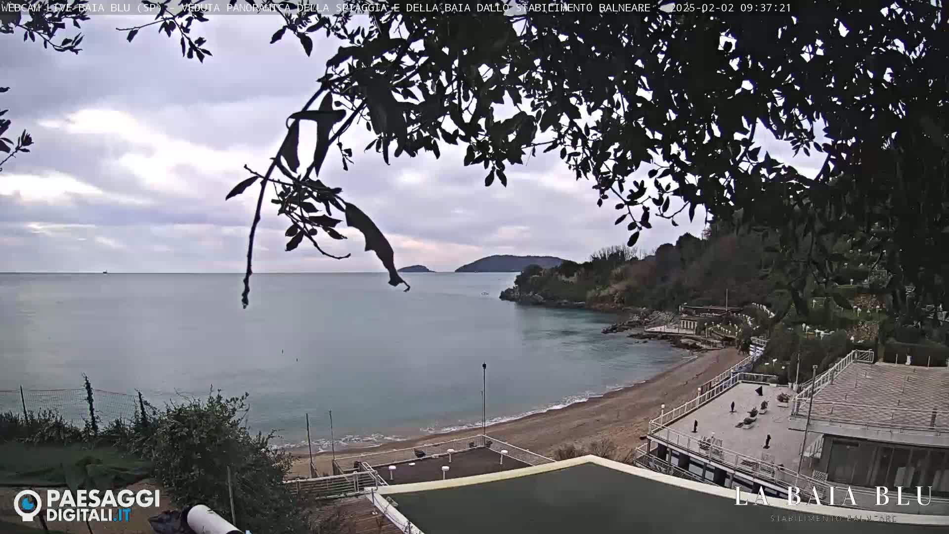 A partly cloudy day shows a sandy beach and calm bay, viewed from a seaside establishment with a few people present.