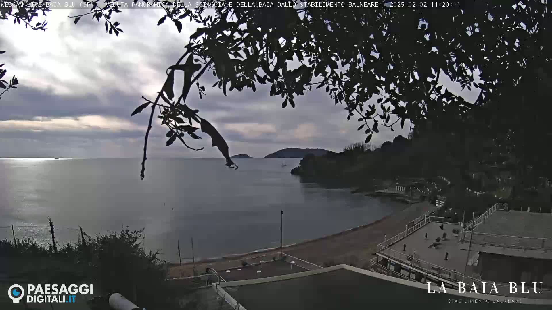A mostly cloudy day overlooks a calm bay with a sandy beach and buildings, partially obscured by tree branches in the foreground.
