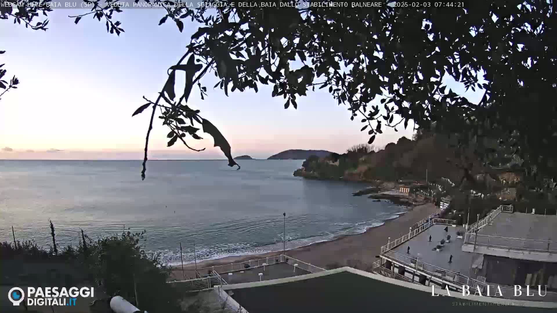 A calm, twilight-lit ocean bay is viewed from above, partially obscured by overhanging tree branches, showing a sandy beach and a seaside establishment.