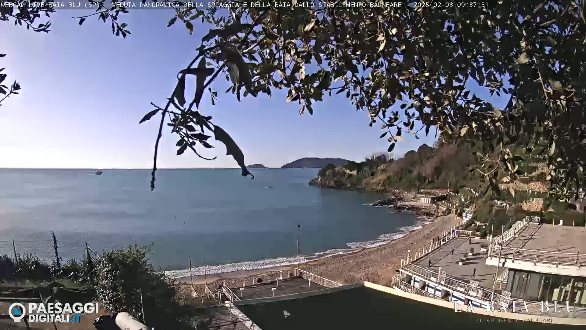 A mostly sunny day overlooking a calm bay with a sandy beach and buildings, partially obscured by tree branches in the foreground.