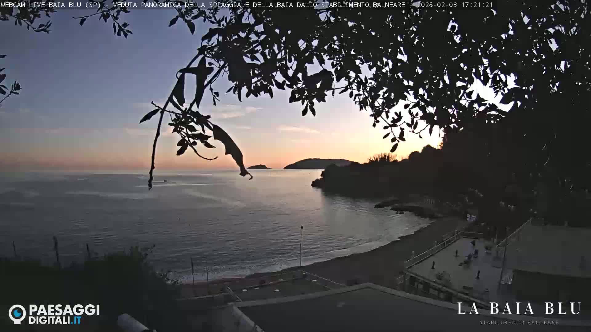 A tranquil beach scene at sunset, viewed through silhouetted tree branches, shows calm water and a sandy shore with a few people present.