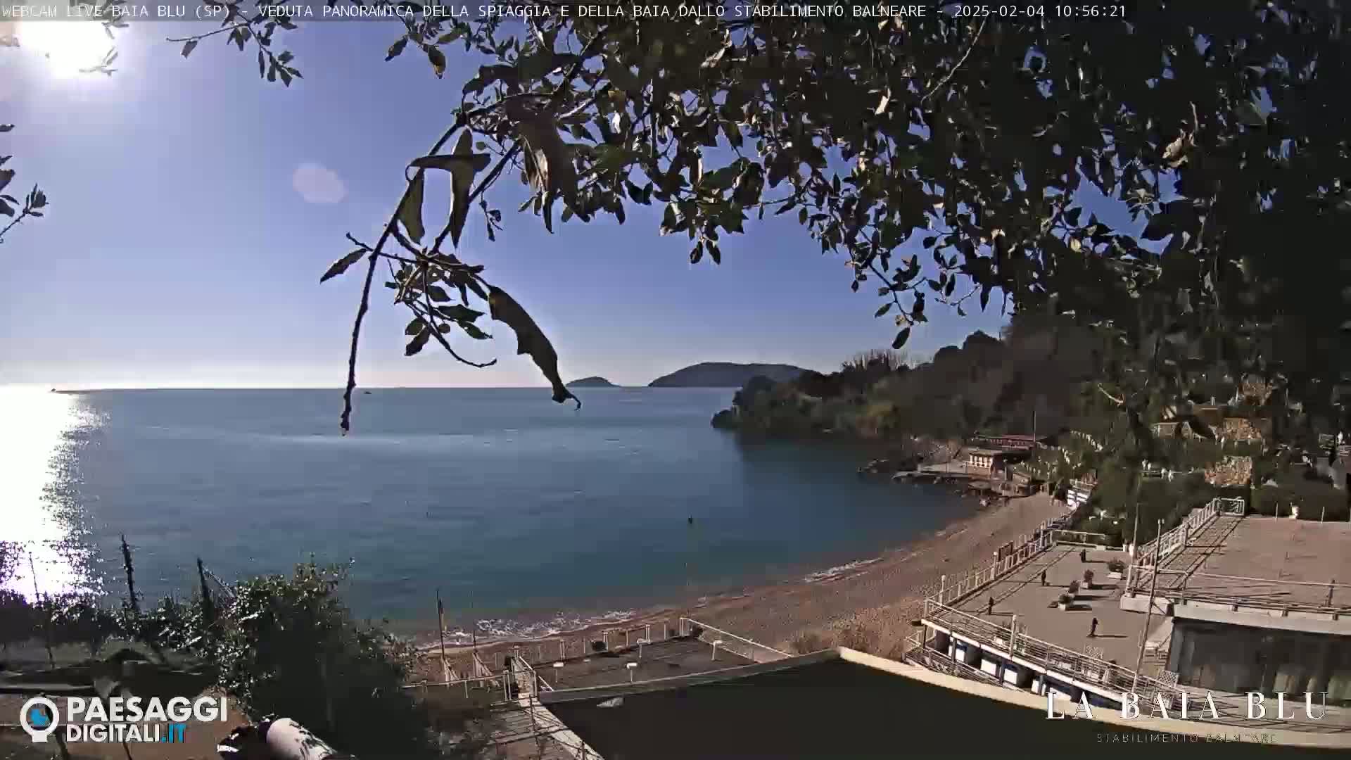 A sunny day overlooking a calm bay with a pebble beach and beachside buildings.