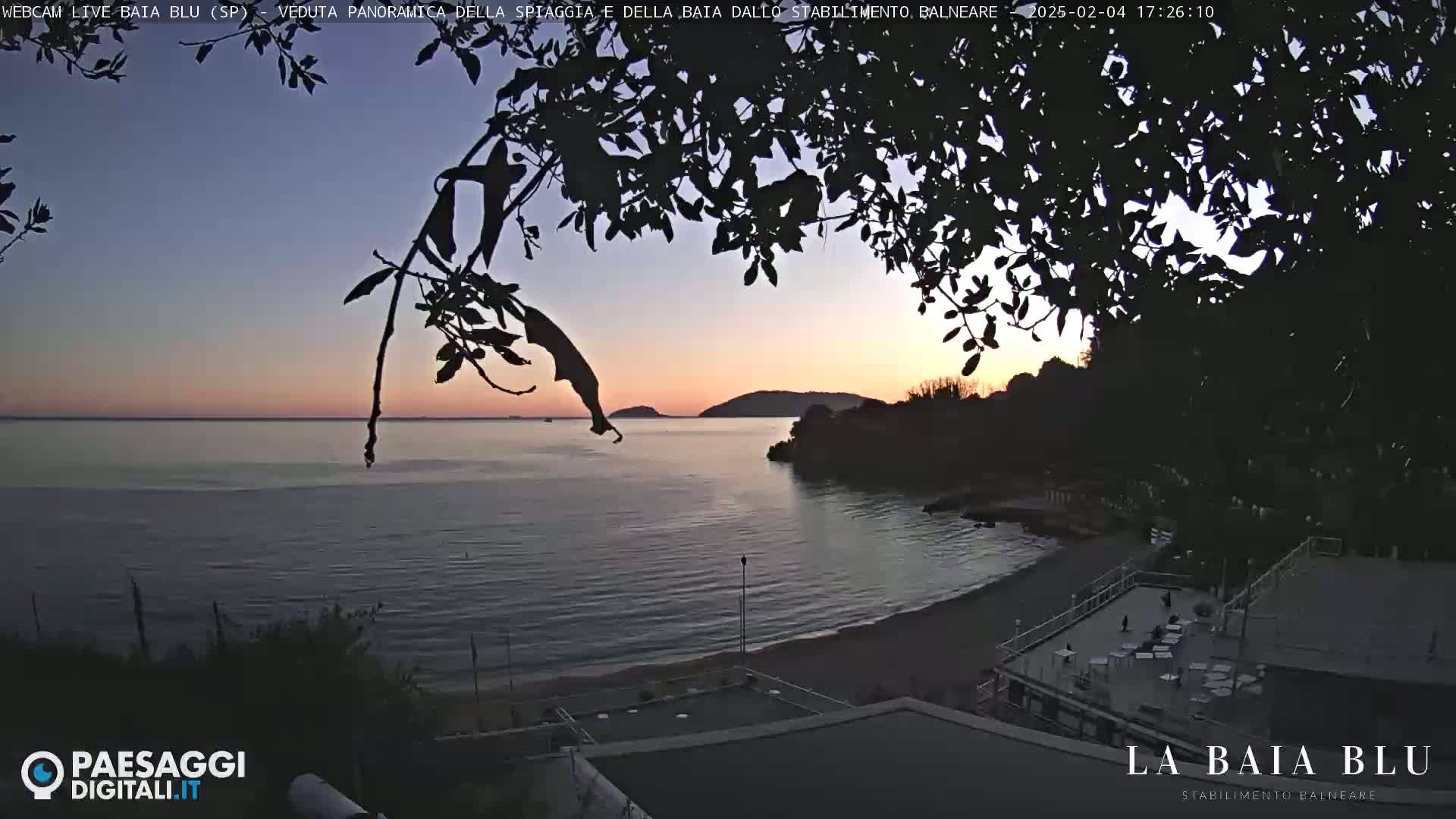 A tranquil beach scene at sunset, viewed from behind silhouetted tree branches, shows calm water and a mostly empty beachside establishment.