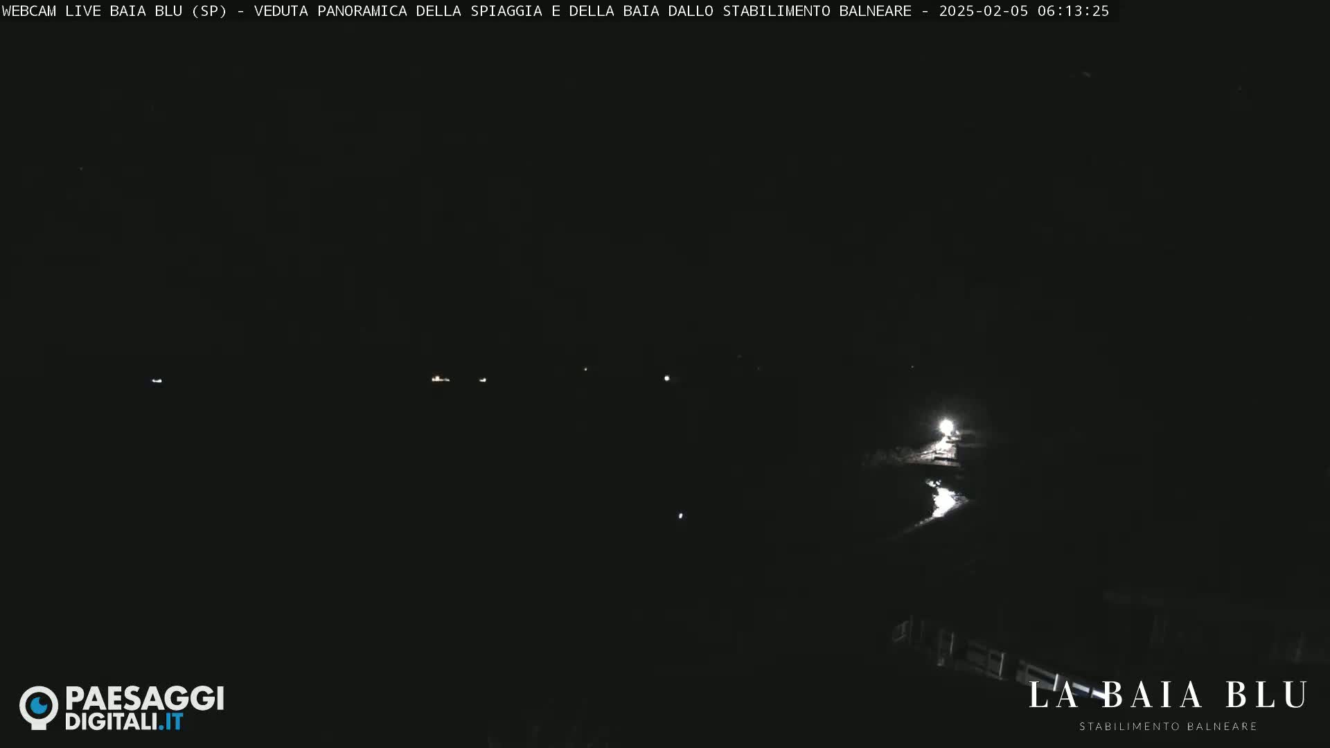 A nighttime panoramic view of a dark beach and bay with a few faint lights visible in the distance.