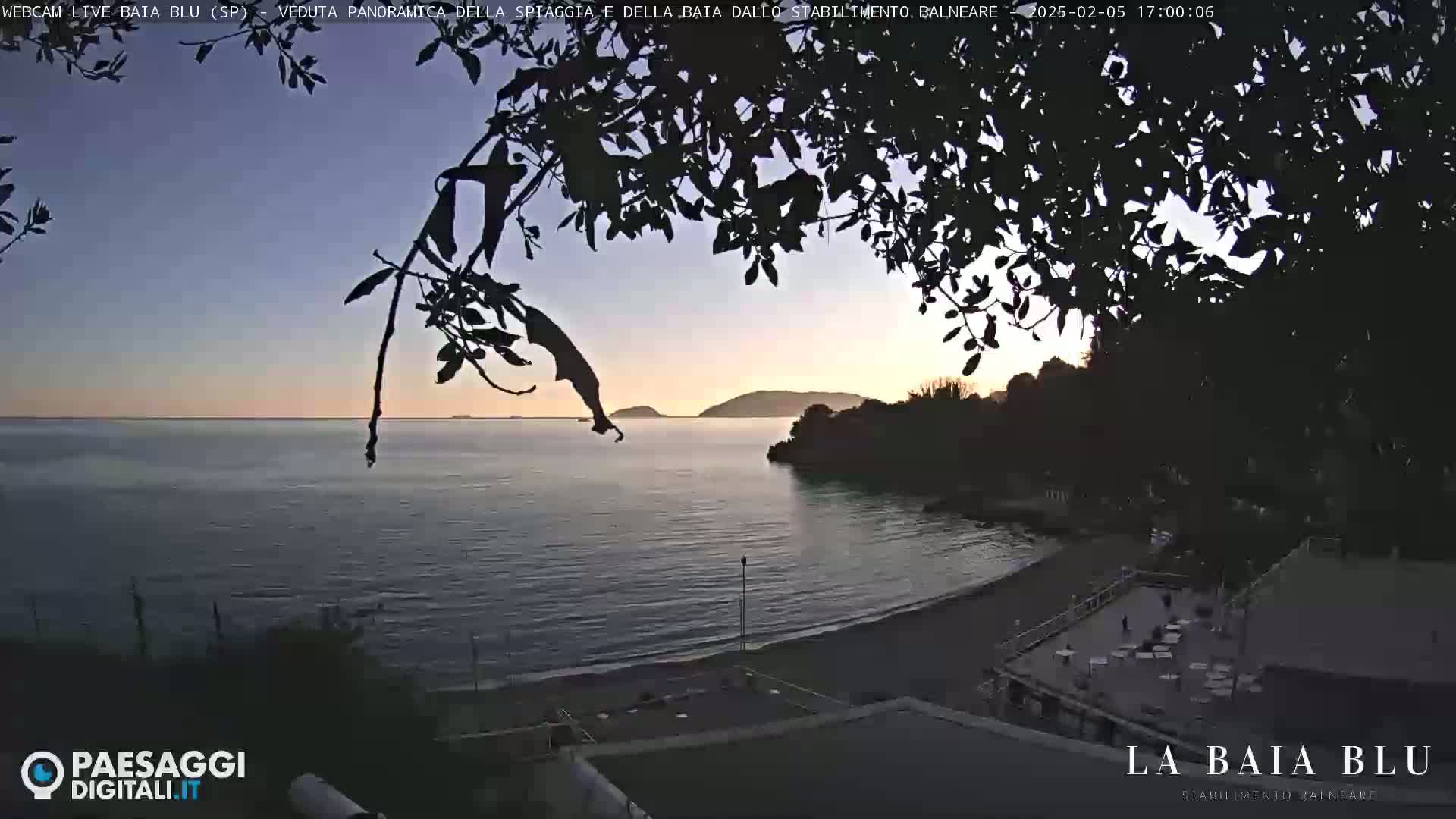A tranquil beach scene at sunset is partially obscured by overhanging tree branches, showing a calm body of water with a distant shoreline.