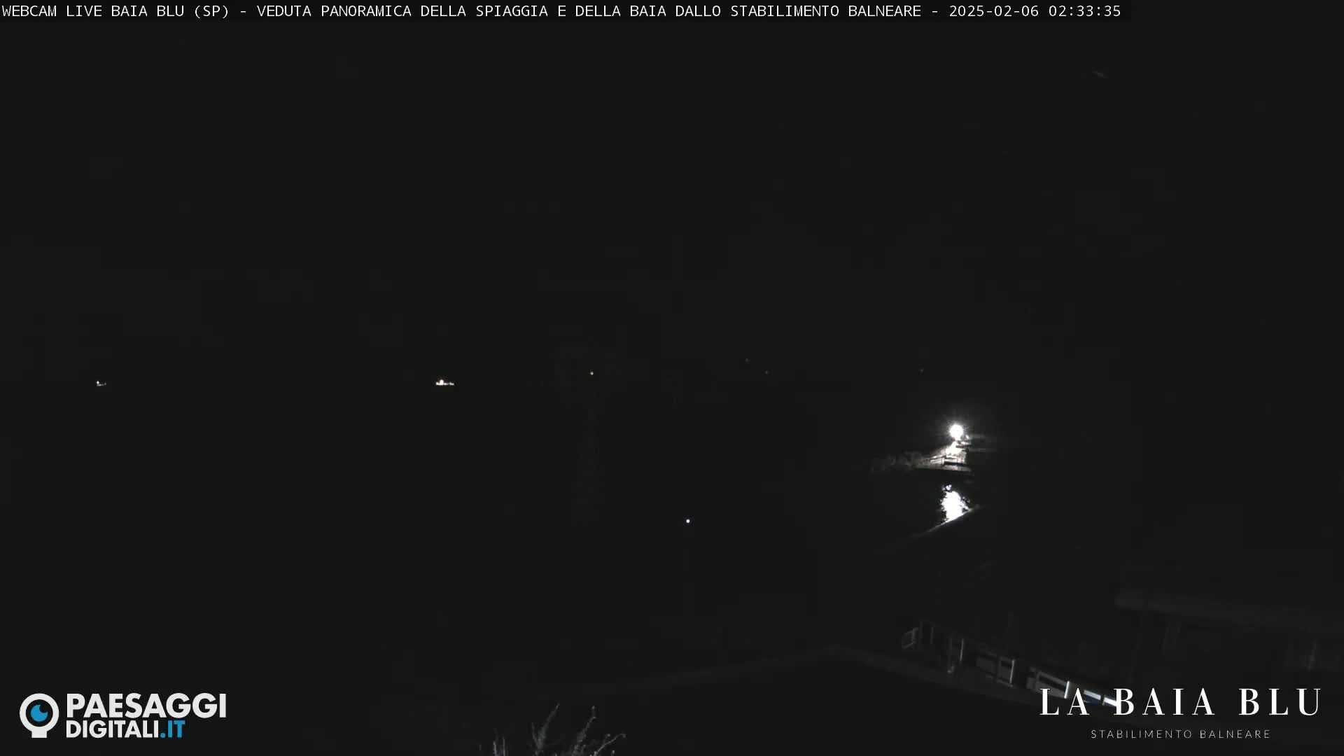 A dark, nighttime view of a beach and bay, with a few faint lights visible in the distance.