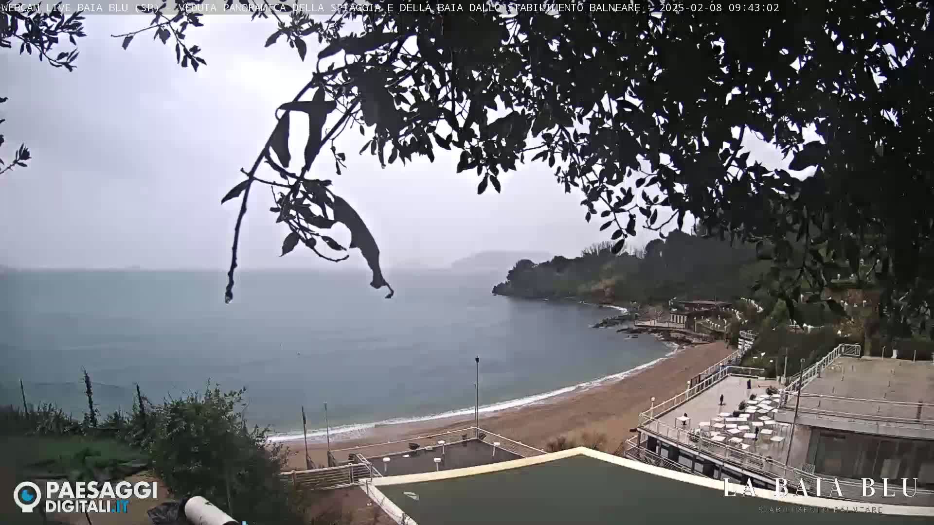 A view from above shows a beach with a building and some trees, overlooking a calm bay on an overcast day.