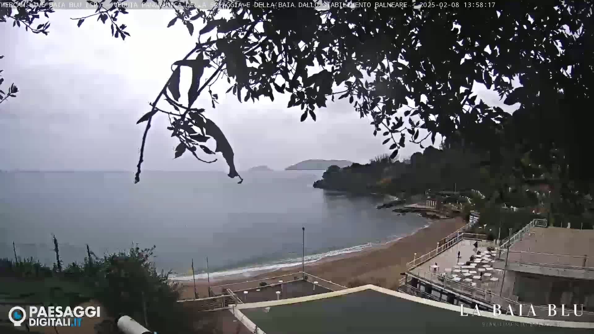 A seaside view on an overcast day shows a sandy beach, calm water, and a partially obscured building with outdoor seating.