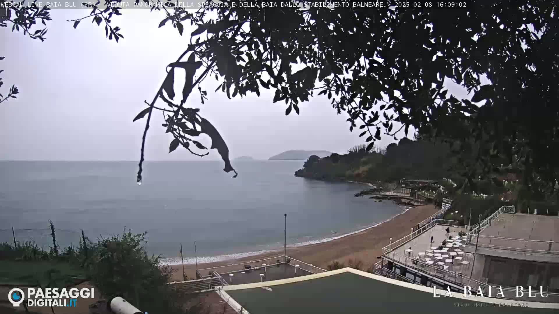 A rainy, overcast day at a beach with calm water, a sandy shore, and a building with outdoor seating overlooking the scene.