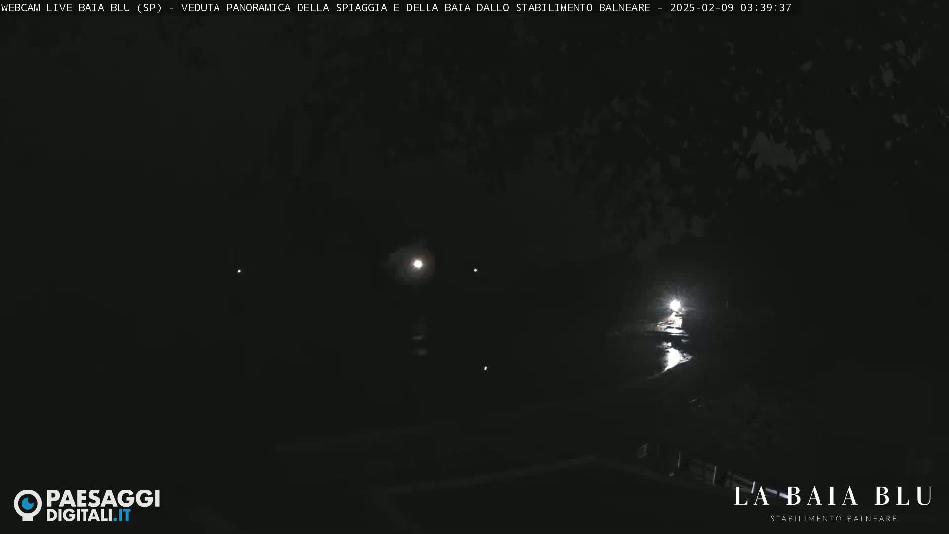 A dark nighttime scene shows a dimly lit body of water reflecting lights, with dark land or trees in the background.