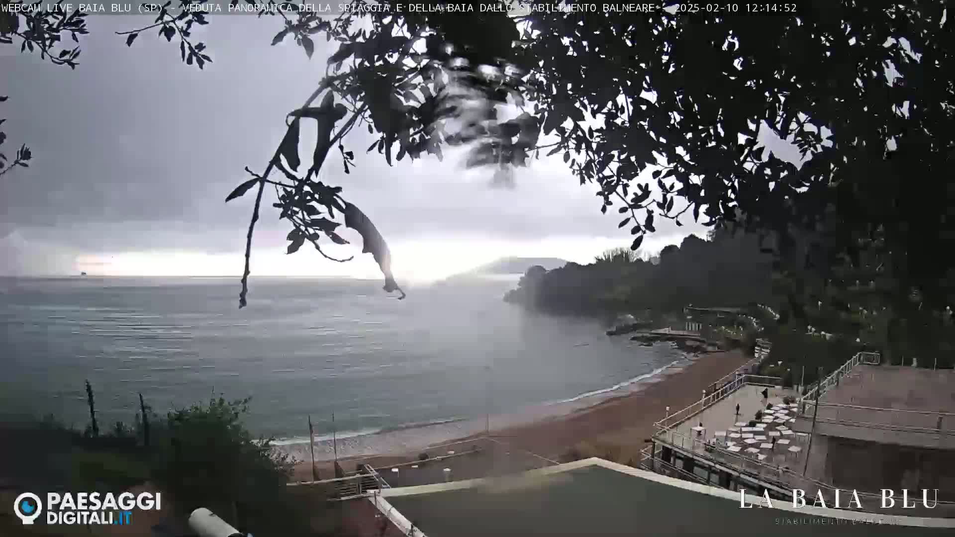A mostly cloudy day shows a view of a beach with calm water and a seaside restaurant, partially obscured by tree branches.