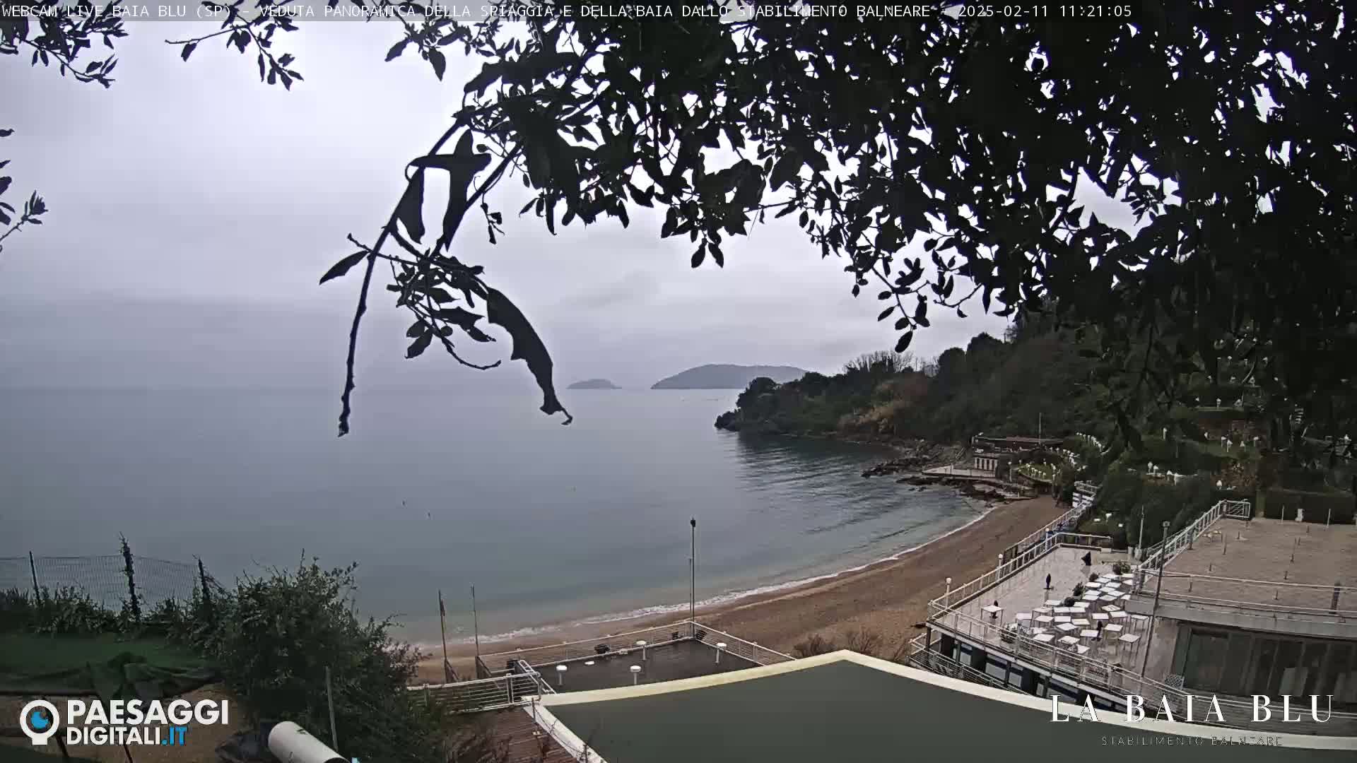 An overcast day shows a sandy beach curving into a calm bay, with dark green hills and a few buildings visible in the background.