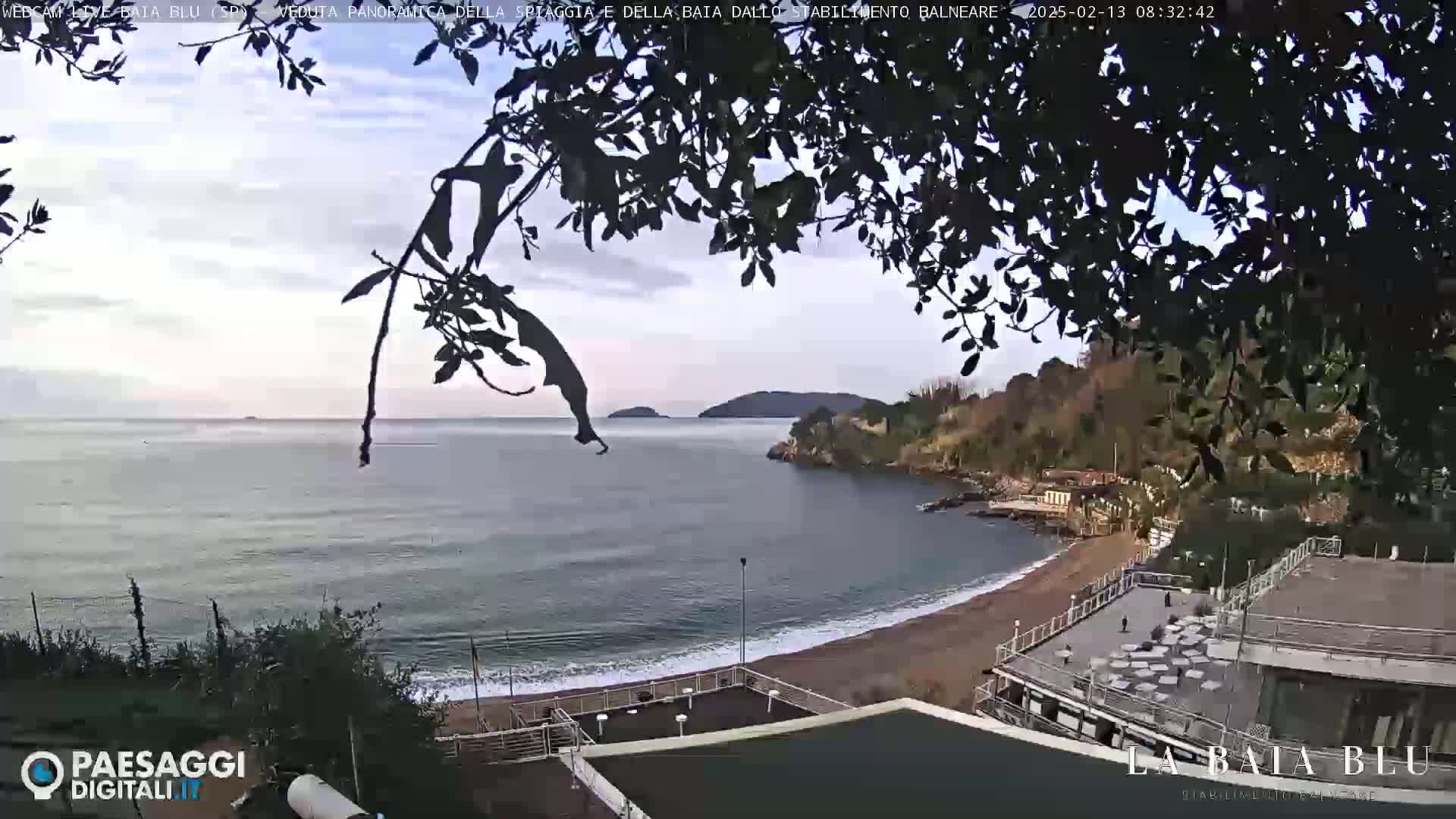 A mostly cloudy day overlooks a calm bay with a sandy beach and buildings, partially obscured by tree branches.
