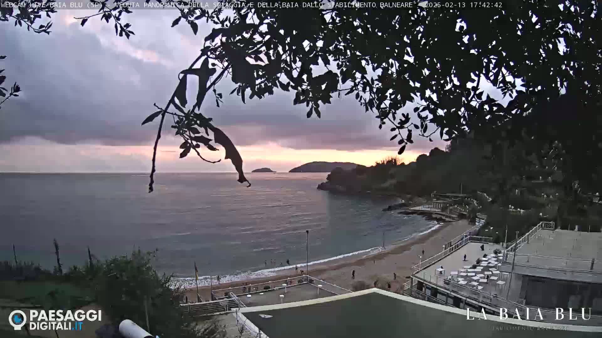 An overcast sunset over a calm bay with a sandy beach and beachside buildings.