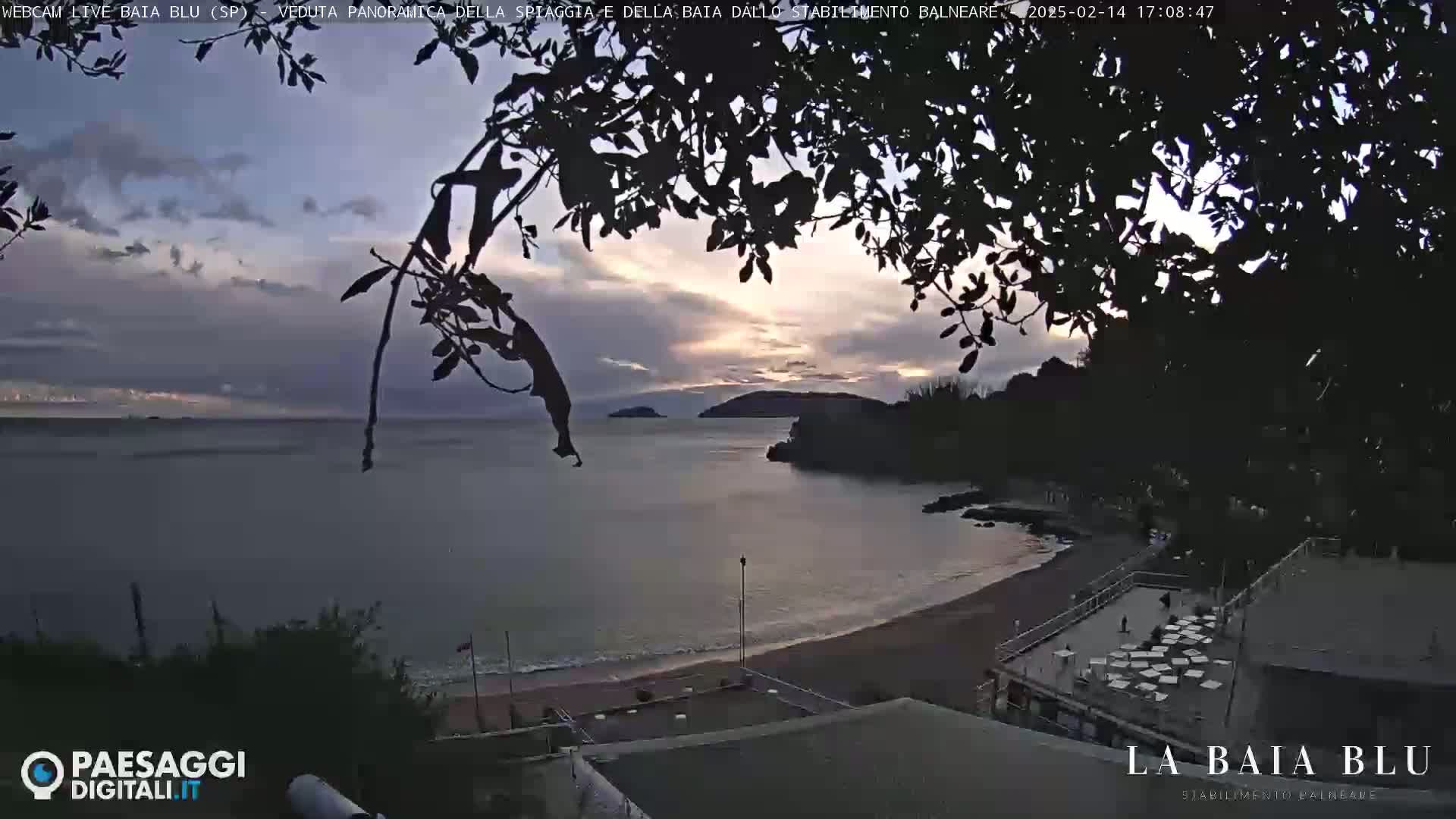 A partly cloudy sunset over a calm bay with a sandy beach and beachside structures is seen through silhouetted tree branches.