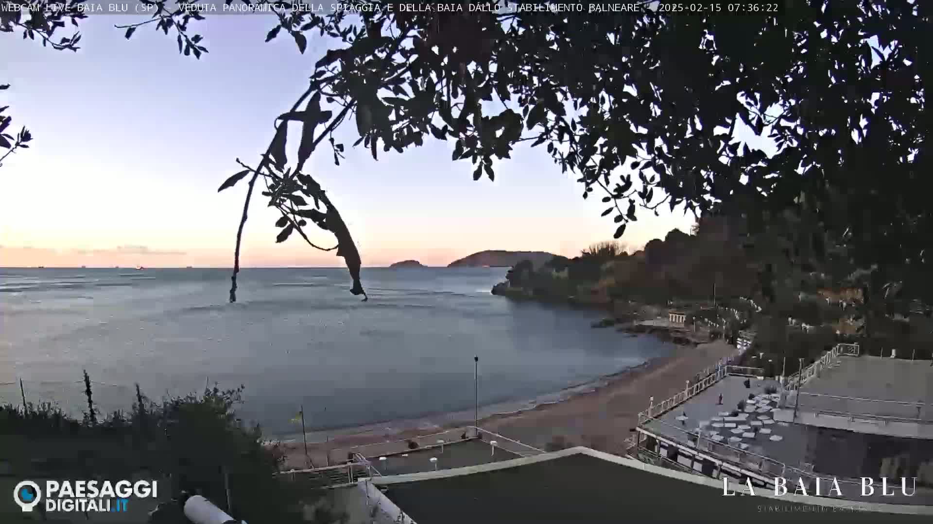 A tranquil beach scene at dawn, with calm water and a few boats visible in the distance, viewed through silhouetted tree branches.