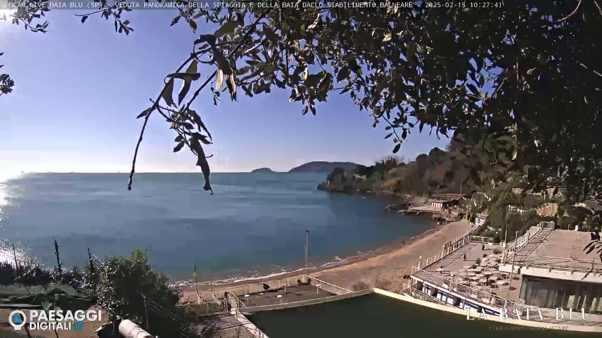 A sunny, clear day overlooking a calm bay with a sandy beach and seaside buildings partially obscured by tree branches.