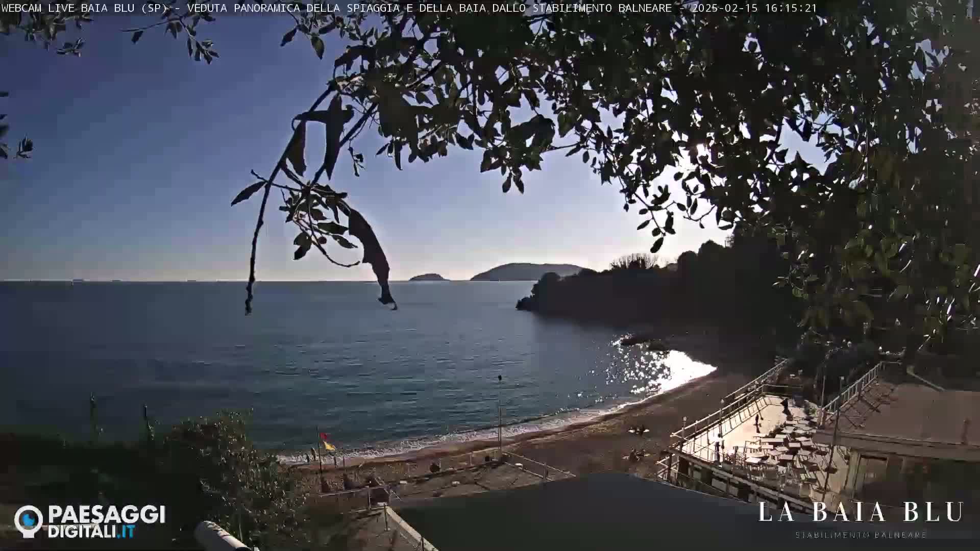 A sunny, partly cloudy day is shown at a beach with calm, blue water, a sandy shore, and a seaside establishment partially obscured by tree branches.