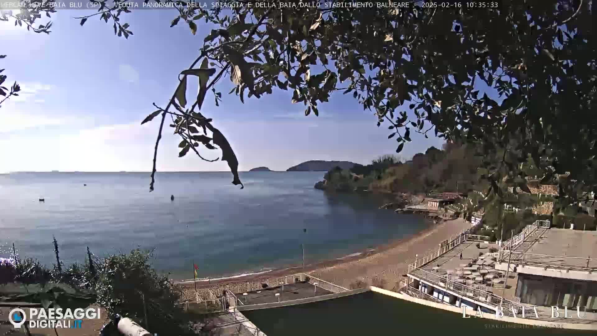 A partly sunny day shows a view of a calm bay with a sandy beach and buildings, partially obscured by tree branches.