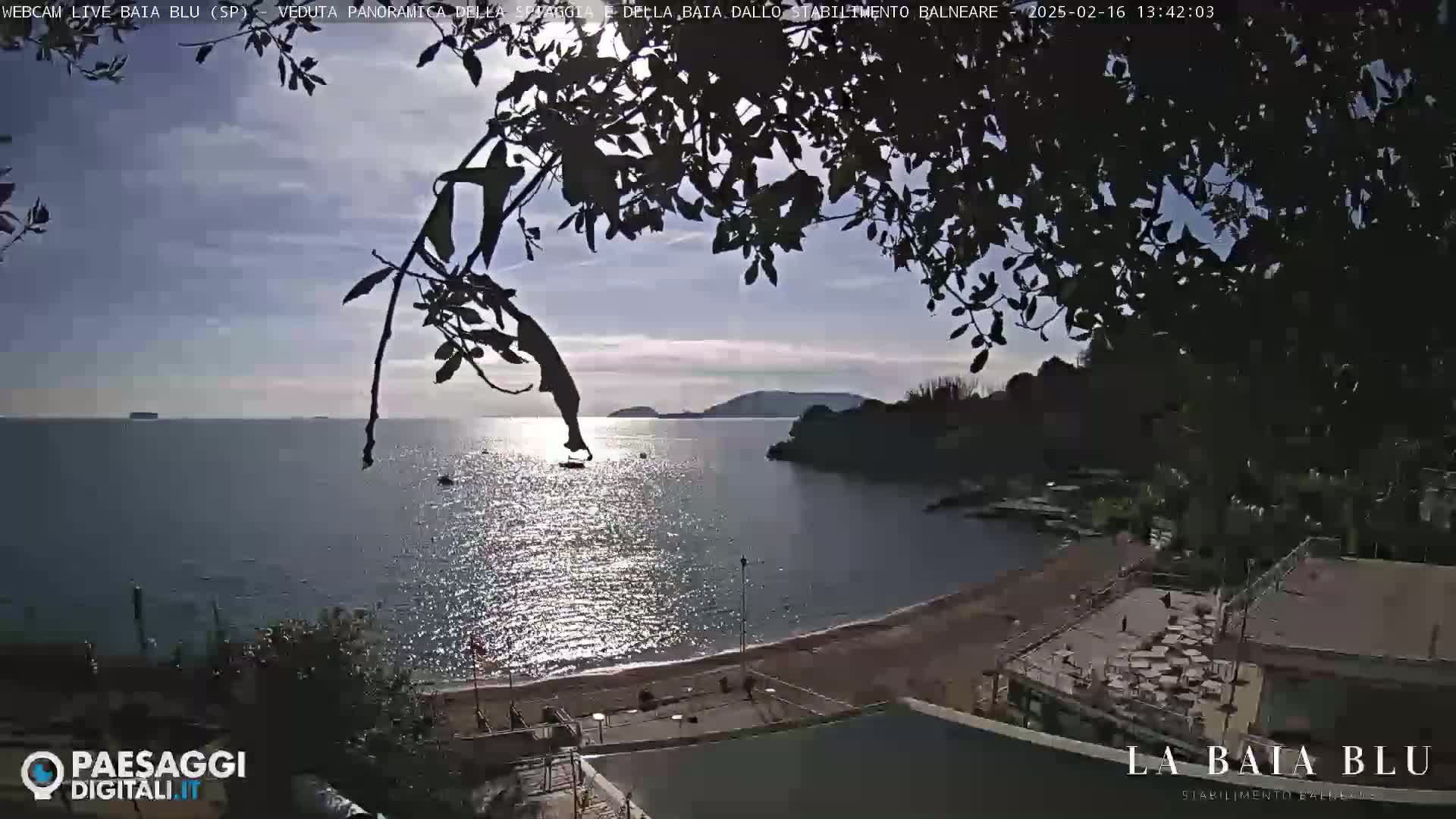 A sunny, partly cloudy day overlooks a calm bay with a sandy beach and seaside establishments, partially obscured by overhanging tree branches.