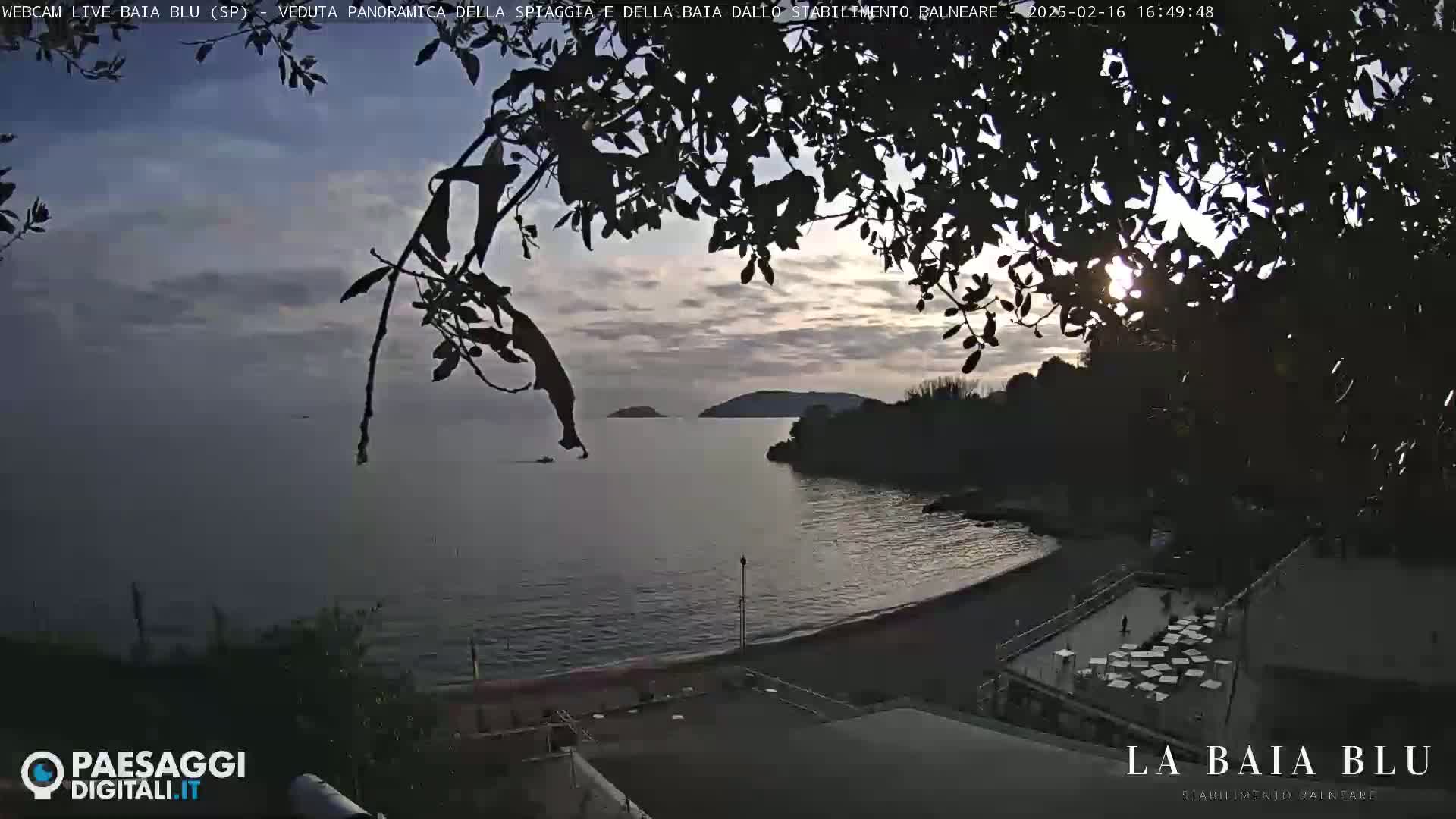 A partly cloudy sunset over a calm bay with a sandy beach and a seaside establishment viewed through silhouetted tree branches.