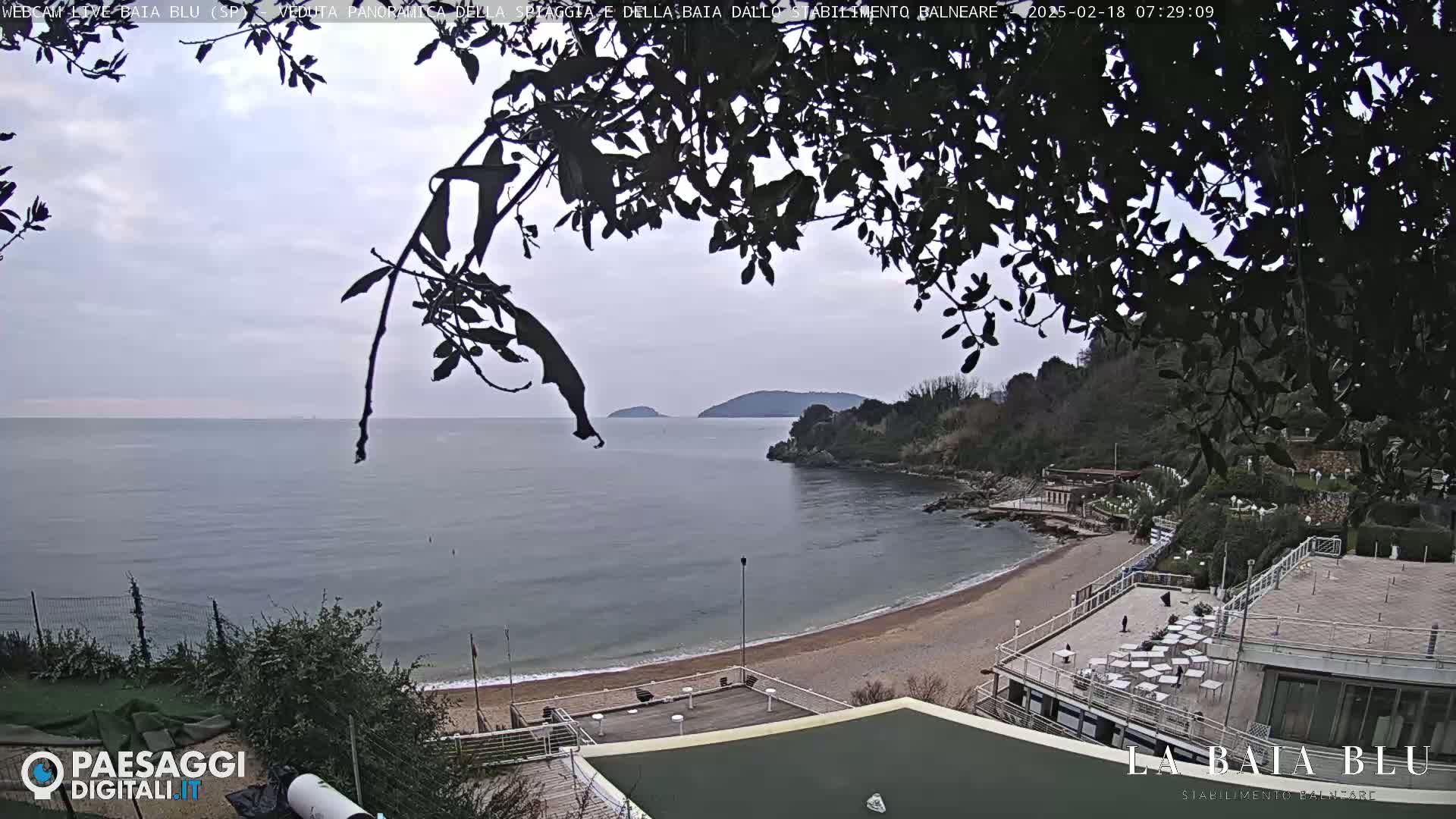 A mostly cloudy day reveals a beach with a building and a calm bay, partially obscured by tree branches, with hills and islands in the distance.