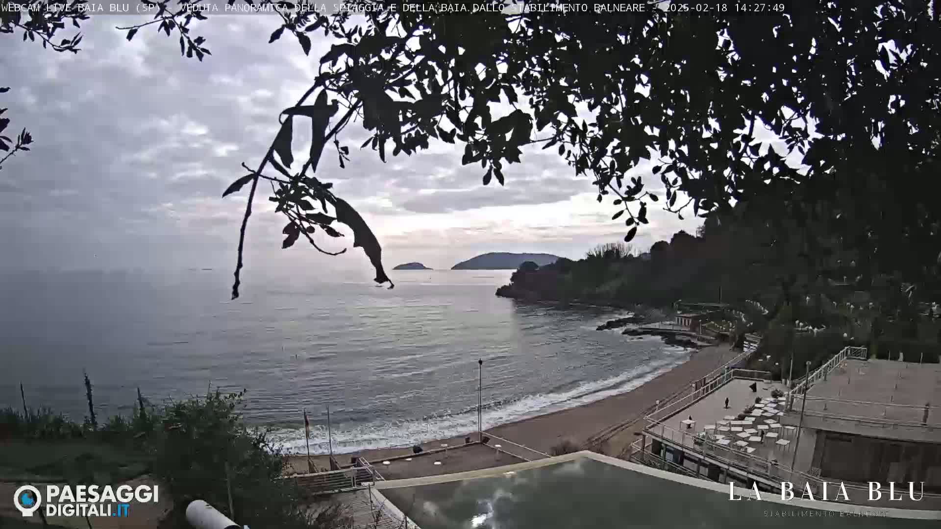 An overcast sky hangs over a sandy beach and calm sea, with distant islands visible and silhouetted trees partially obscuring the view from an elevated vantage point.