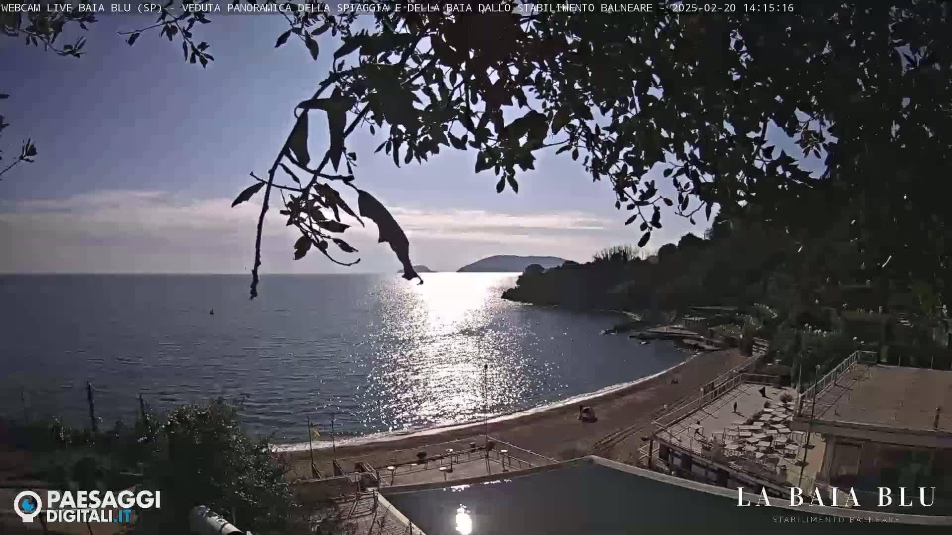 A sunny day overlooking a calm bay with a sandy beach and seaside establishment, partially obscured by silhouetted tree branches.