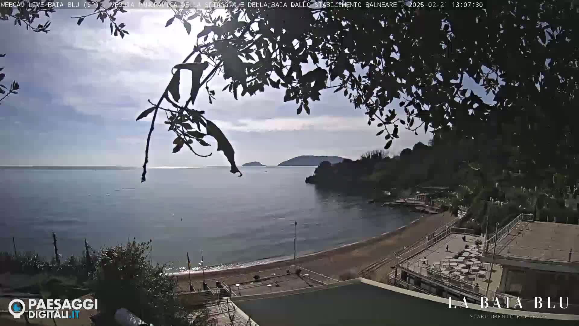 A partly sunny day shows a sandy beach and calm bay with a small landmass in the distance, partially obscured by tree branches in the foreground.
