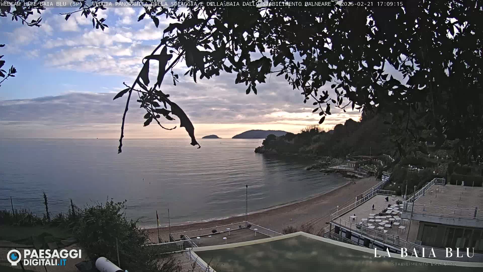 A partly cloudy sunset over a calm bay with a sandy beach and buildings visible from behind a silhouetted tree.