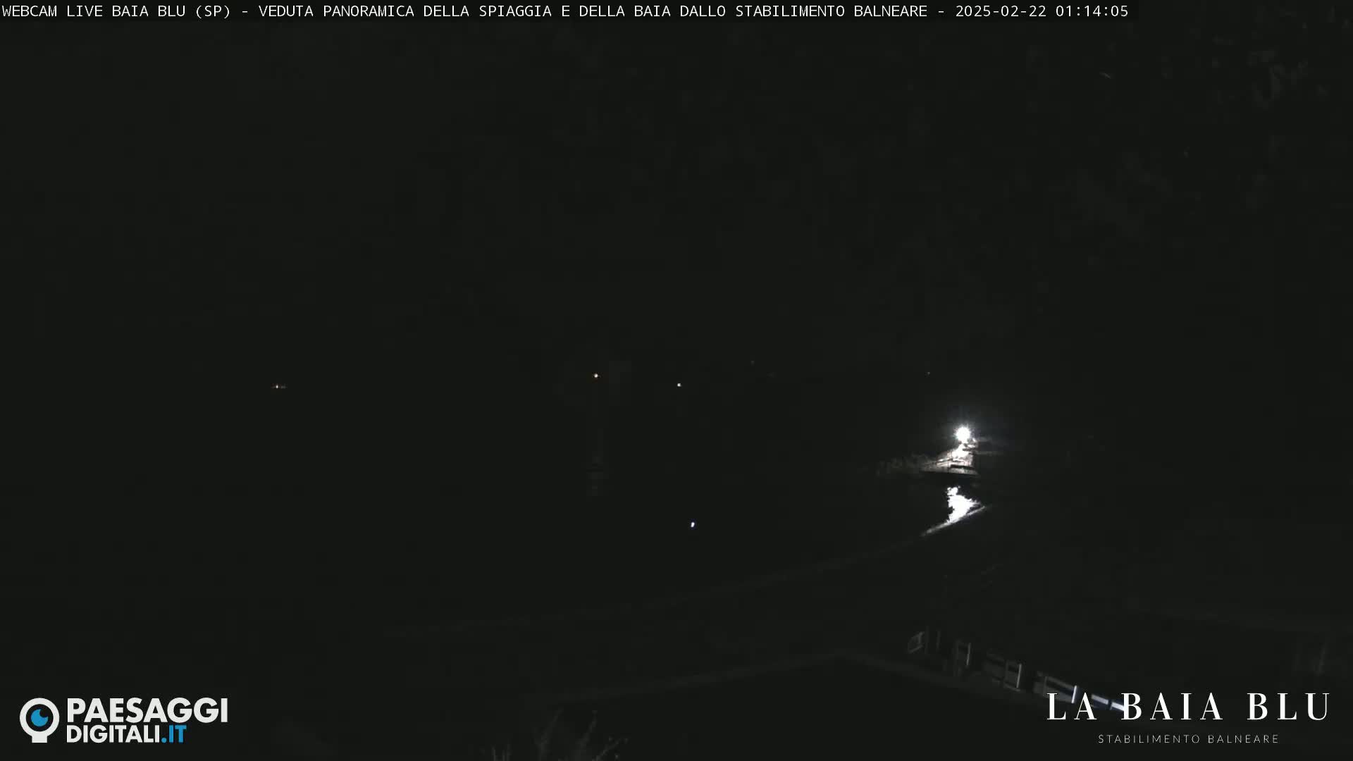 A nighttime, dark view of a beach and bay with a single bright light source reflecting on the water.